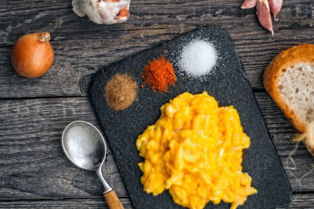 Top view of delicious scrambled eggs served on black board with colorful spices on wooden table with onion and slice of bread
